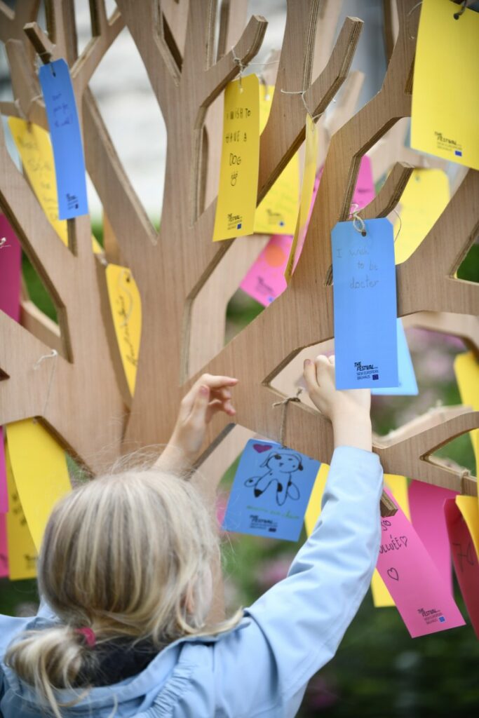 A person hanging a note on the wooden tree installation at the New European Bauhaus 2026